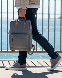 Man walking with a sleek black HP backpack with brown leather straps (Envy Backpack) along a sunny waterfront promenade 