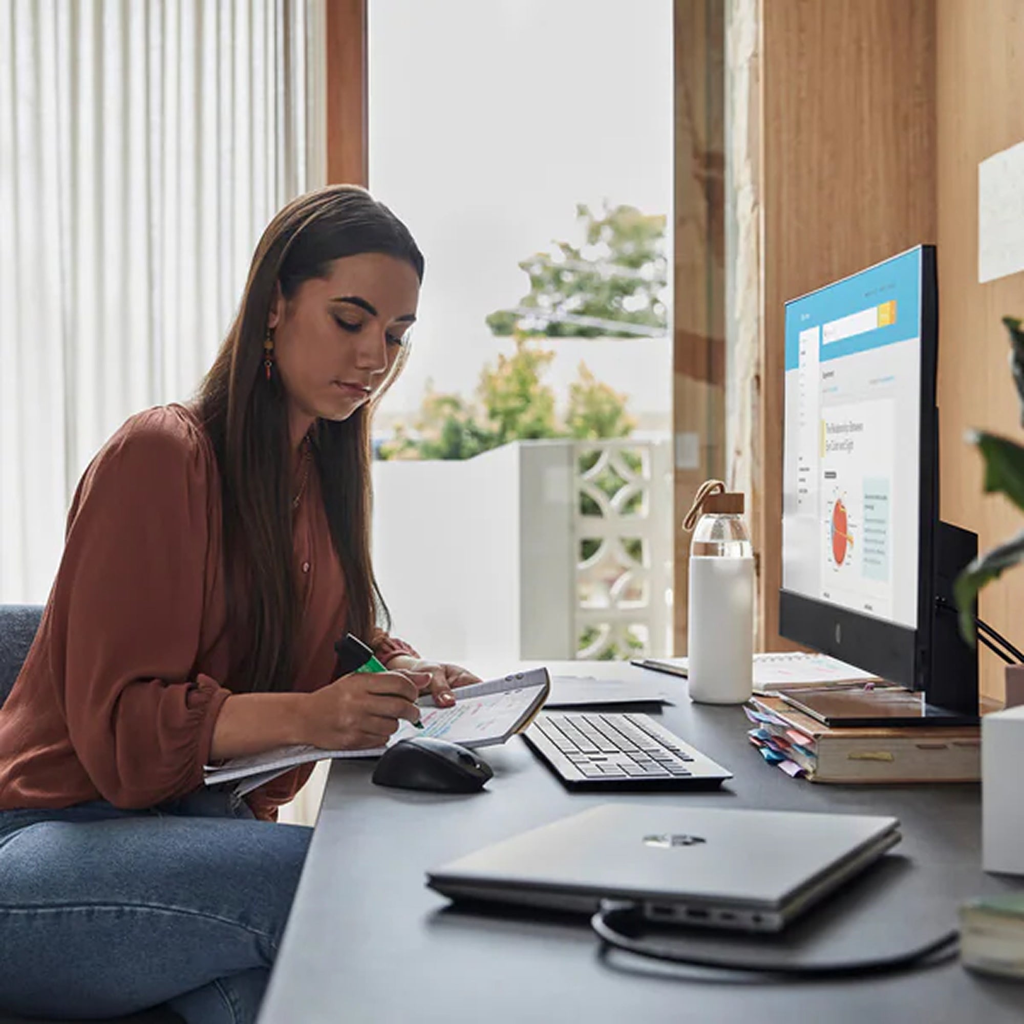 Woman working at a desk with HP All-in-One PC and a notebook in a bright room.